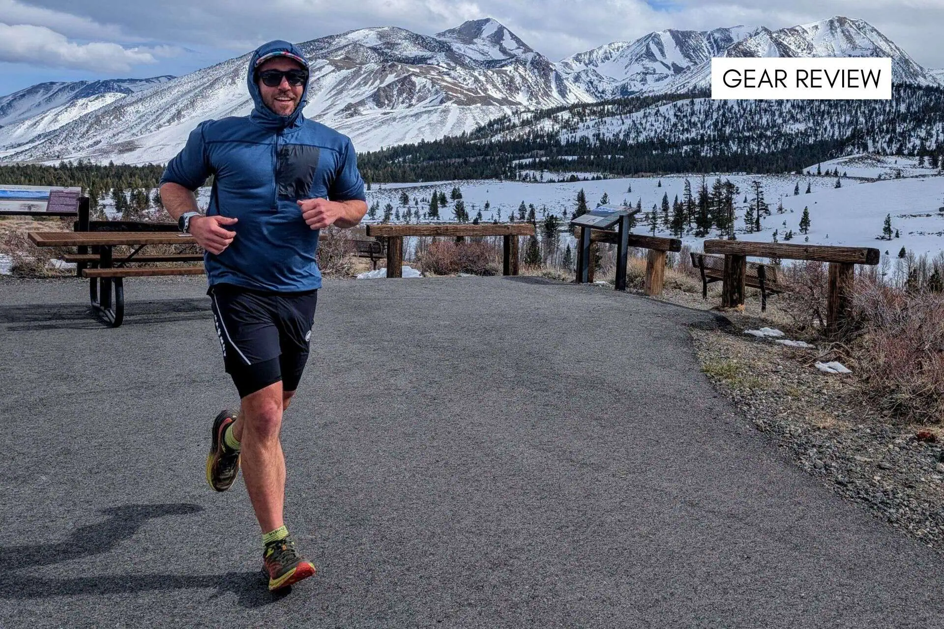 A person in Ridge Merino Solstice Pro athletic gear is jogging on a paved path with snowy mountains and pine trees in the background. A table and benches are on the left. The sky is partly cloudy. Text in the top right reads GEAR REVIEW.