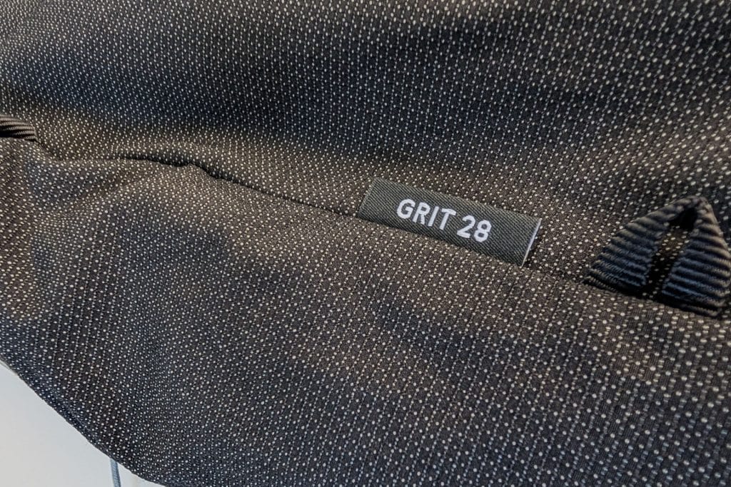 Close-up of a textured black fabric with a small white label reading GRIT 28 attached. The material, part of the Gossamer Gear Grit 28L Fastpack, has a subtle dotted pattern, with two loops visible on the side.