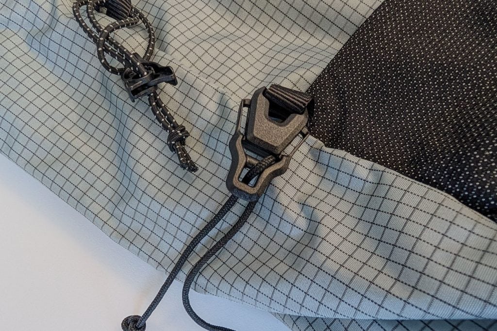 Close-up of a gray grid-patterned fabric on the Gossamer Gear Grit 28L Fastpack, featuring a black adjustable buckle and drawstring. The drawstring boasts a sliding knot, while the fabric is partially black with small white speckles.