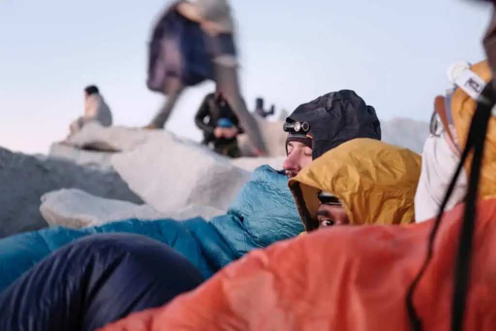 A group of climbers in bright-colored jackets and headlamps huddle together on rocky terrain, capturing one of 2024s best PCT moments. They seem to be resting or sheltering, while someone stands in the background under a clear sky.