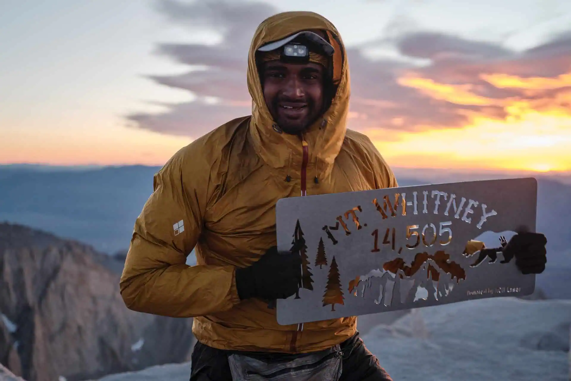 At sunrise, a person in a yellow jacket and headlamp proudly holds a metal sign reading Mt. Whitney 14,505. With the majestic mountains and vibrant sky behind them, it encapsulates one of 2024s best PCT moments.
