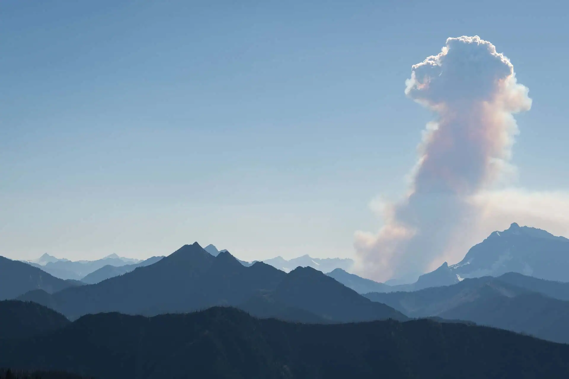A distant view of mountainous terrain with a large plume of smoke rising skyward from a peak on the right captures one of natures worst moments. The sky is clear, with the sun casting shadows over the rugged landscape, reminiscent of pct 2024 forecasts.