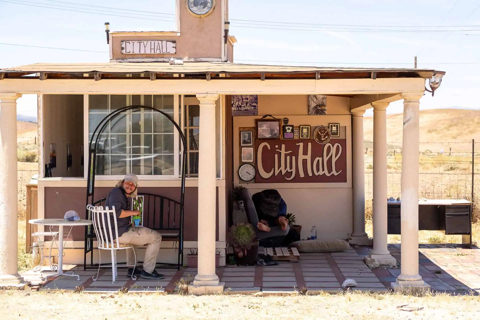 A person sits on a porch swing outside a small, rustic City Hall building, capturing one of the best moments of 2024. The scene features a clock, framed pictures, and charming decor. Nearby, a dog rests as the man at the table enjoys his drink amidst dry grass and open fields.