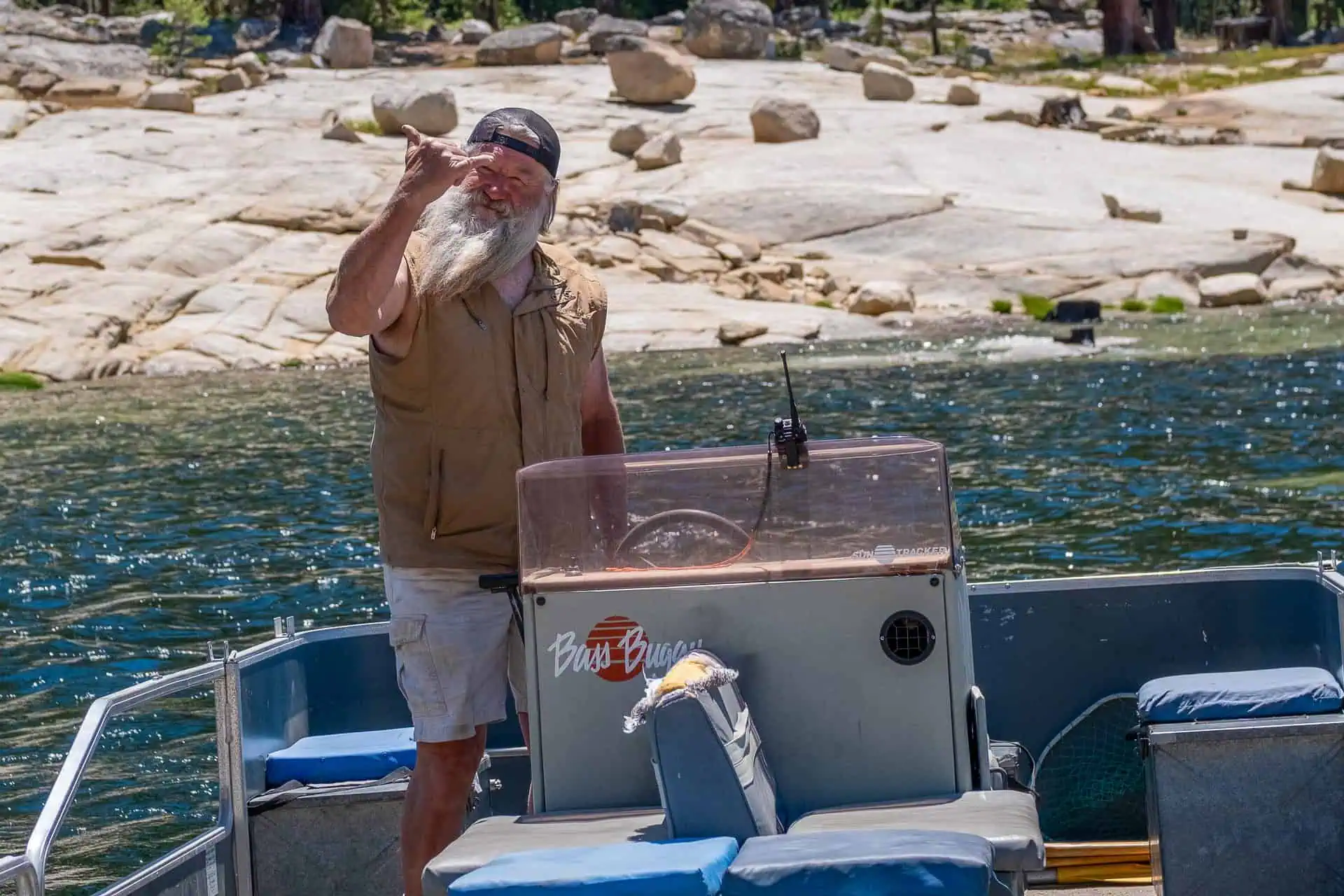A person with a long white beard, wearing a beige vest and shorts, gestures enthusiastically while standing on a small boat. Its one of the best 2024 pct moments, set against rocky shores and a clear sky over the lake.