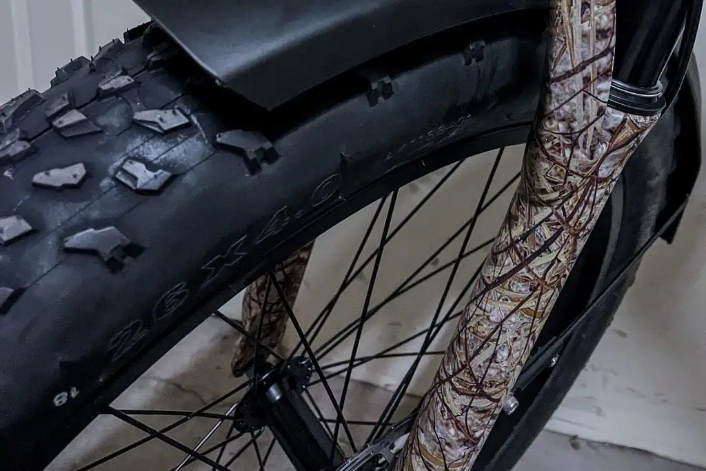 Close-up of a PUCKIPUPPY Bulldog bikes front wheel with a large, knobby 26 x 4.0 tire, attached to a fork featuring a brown, camouflage-like pattern. The bike is indoors against a light-colored wall.