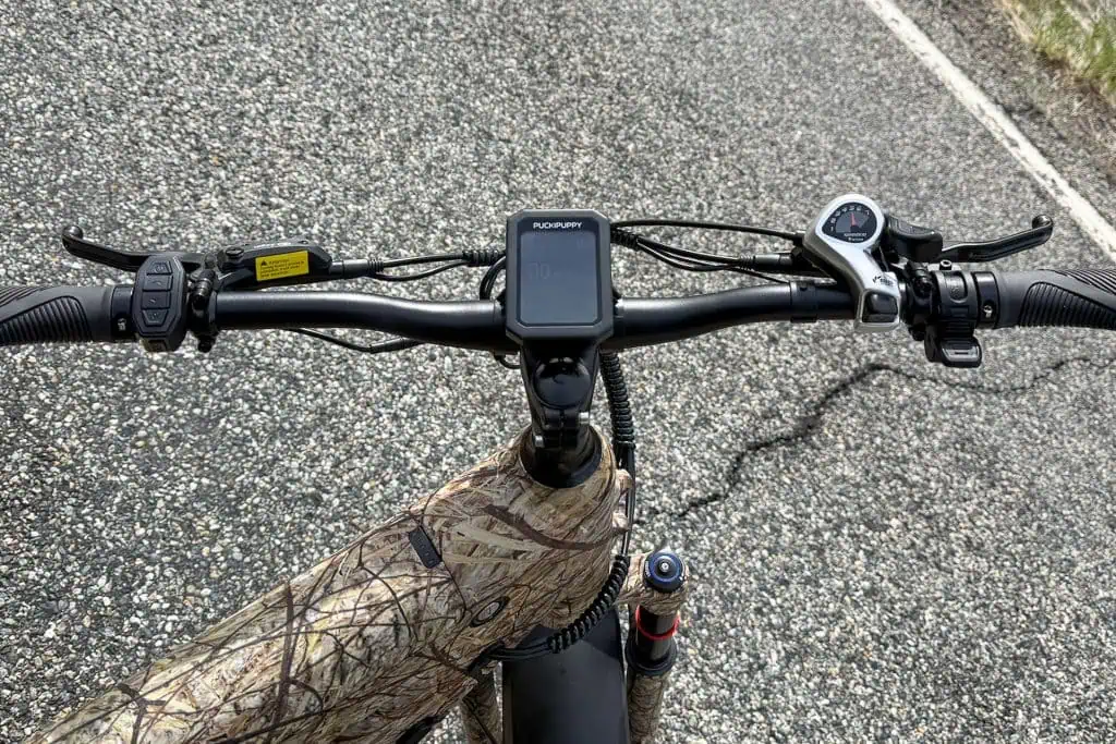 Close-up view of a PUCKIPUPPY Bulldog bicycle’s handlebars on a paved road. The camo-patterned frame, screen, buttons, and gear shifter stand out as the slightly rough surface stretches ahead.
