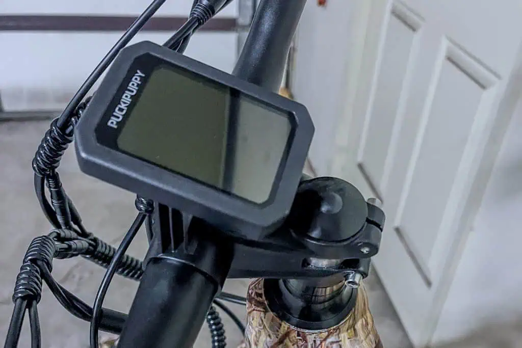 Close-up of a bicycle handlebar featuring a PUCKIPUPPY Bulldog electronic display, with visible wiring and part of a patterned frame, indoors near a white door.