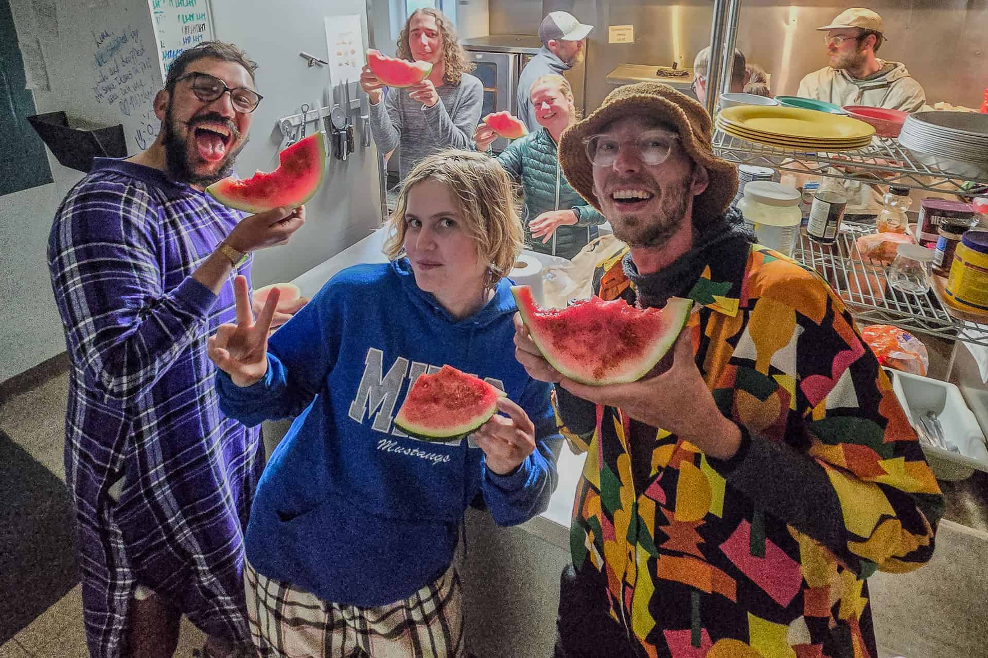 A group of people in a kitchen enjoy slices of watermelon, smiling and posing for the camera. Some wear colorful clothes and hats, one flashes a peace sign—capturing fun, not 2024 CDT worst moments—while others prepare food in the background.
