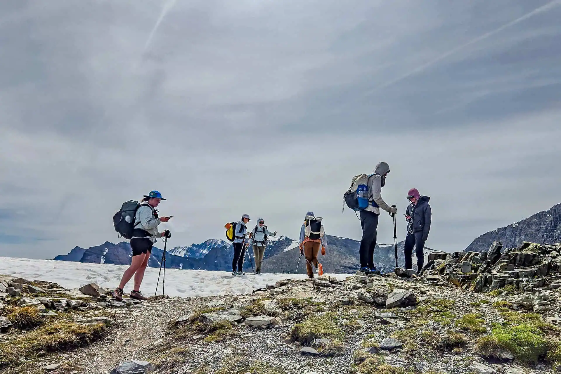 A group of six hikers with backpacks and trekking poles stand on a rocky mountain trail, recalling 2024 CDT worst moments, as snow patches and distant snowy peaks stretch under a cloudy sky.