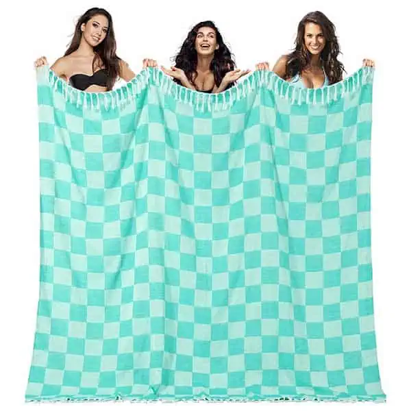 Three women stand behind and hold up a large, turquoise and white checkered blanket with tasseled edges, smiling at the camera—perhaps sharing tips on the best couples sleeping setup for river rafting.