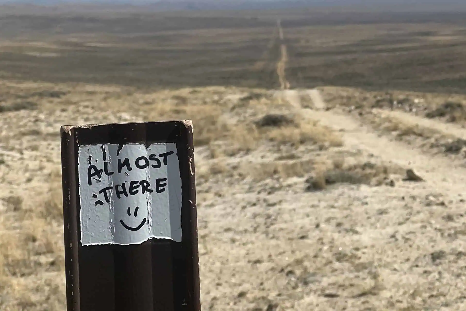 A close-up of a post with a hand-written sign saying Almost there :). In the background, a long dirt road—like the 2024 CDT worst moments—stretches into a dry, flat, and barren landscape under a hazy sky.