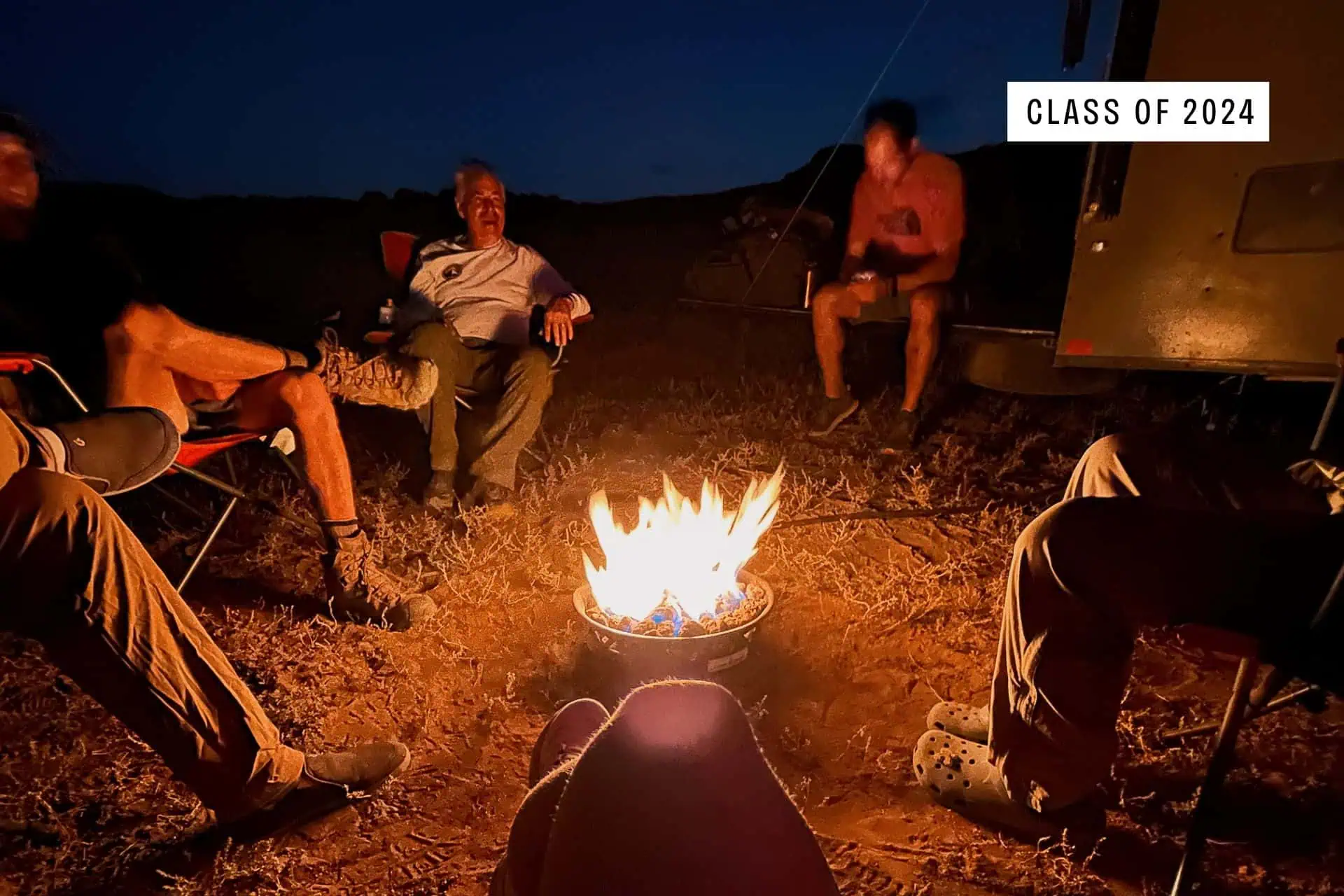Four people sit in a circle around a campfire at night, illuminated by the fire’s glow. The words “Class of 2024” appear at the top right as they share stories about their 2024 CDT worst moments. Camping gear and a trailer are visible in the background.