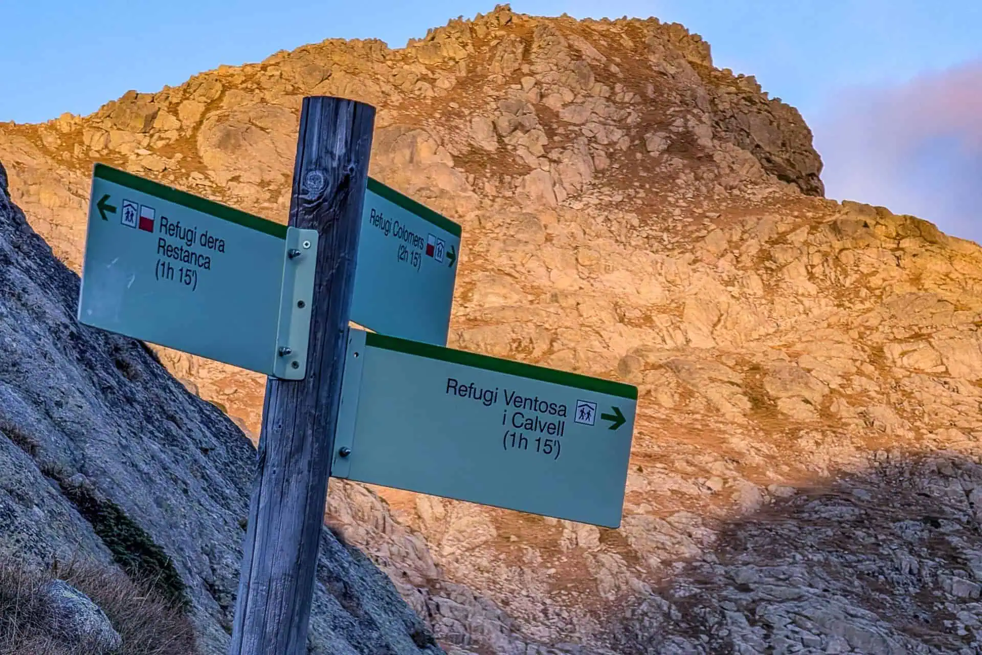 A wooden post with directional hiking signs in Italian points to three mountain refuges along the HRP, set against a rocky, sunlit mountain backdrop. The signs indicate hiking times ranging from 1 hour to 1 hour 15 minutes.
