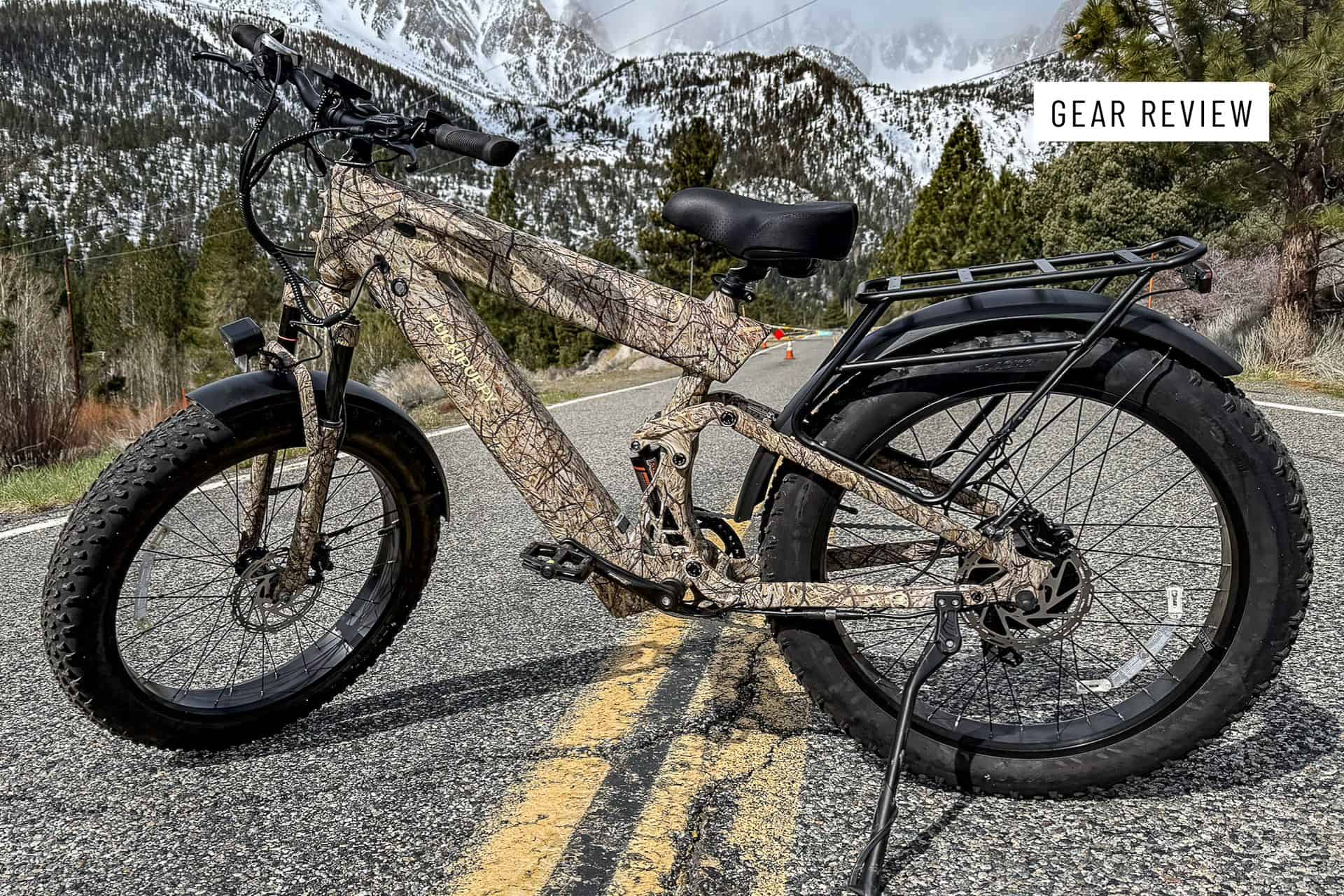 A PUCKIPUPPY Bulldog camouflage-patterned electric fat-tire bike is parked on a mountain road, with snowy peaks and pine trees in the background. The image includes a label reading Gear Review in the top right corner.