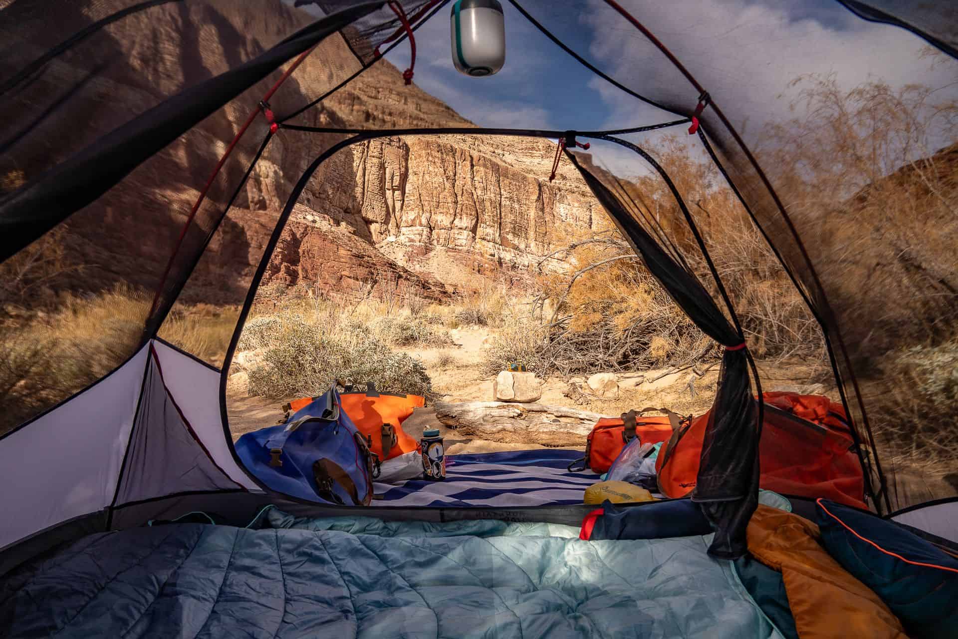 View from inside a tent looking out at a rocky canyon landscape, with a two-person sleeping setup and camping gear scattered on a blanket outside under a partly cloudy sky—perfect for river rafting adventures.