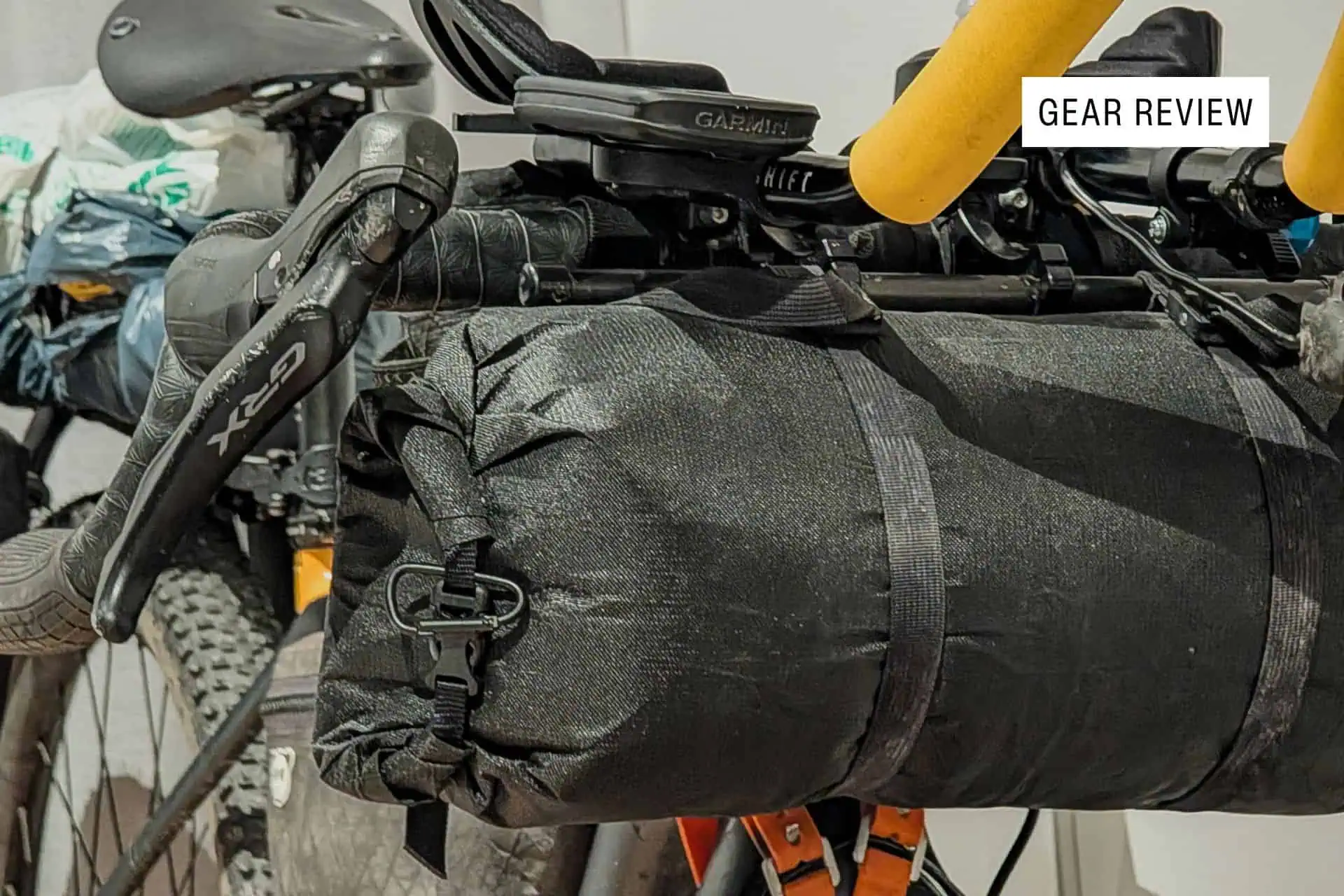 Close-up of a bicycle’s handlebars and front bag, featuring bright yellow grips, bikepacking gear, and the Gossamer Gear Grit 28 pack attached. A label in the corner reads GEAR REVIEW.