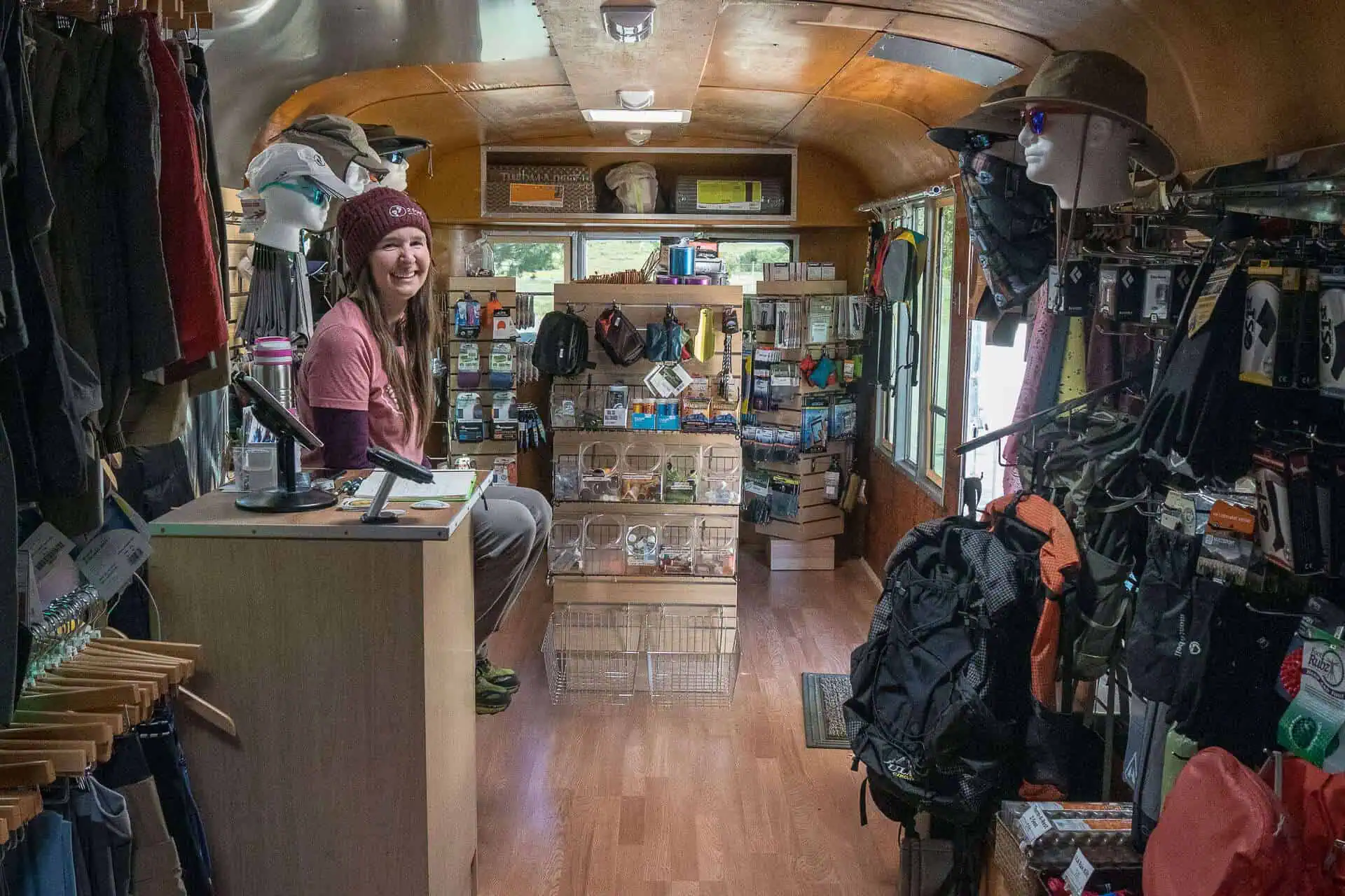 A woman sits smiling behind a counter inside a cozy, well-organized mobile shop filled with outdoor gear, clothing, backpacks, and accessories—ready for anyone chasing their own 2024 best PCT moments.