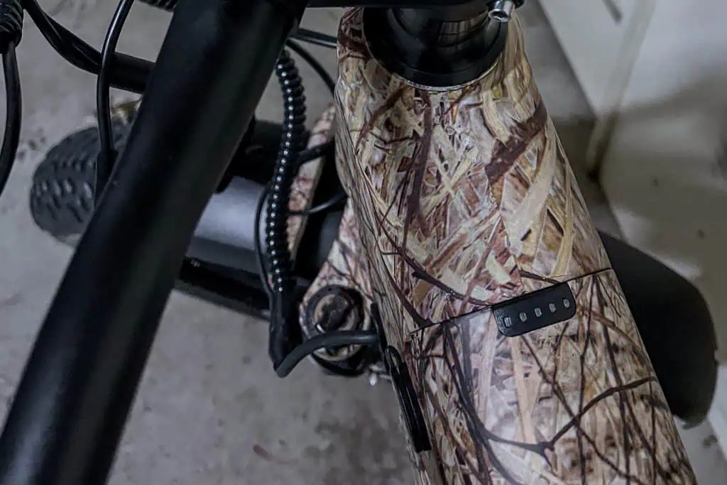 Close-up of a PUCKIPUPPY Bulldog e-bike frame with a camouflage pattern, showing part of the handlebar, cables, and a small LED battery indicator on the top tube. The bike sits indoors on a concrete floor.