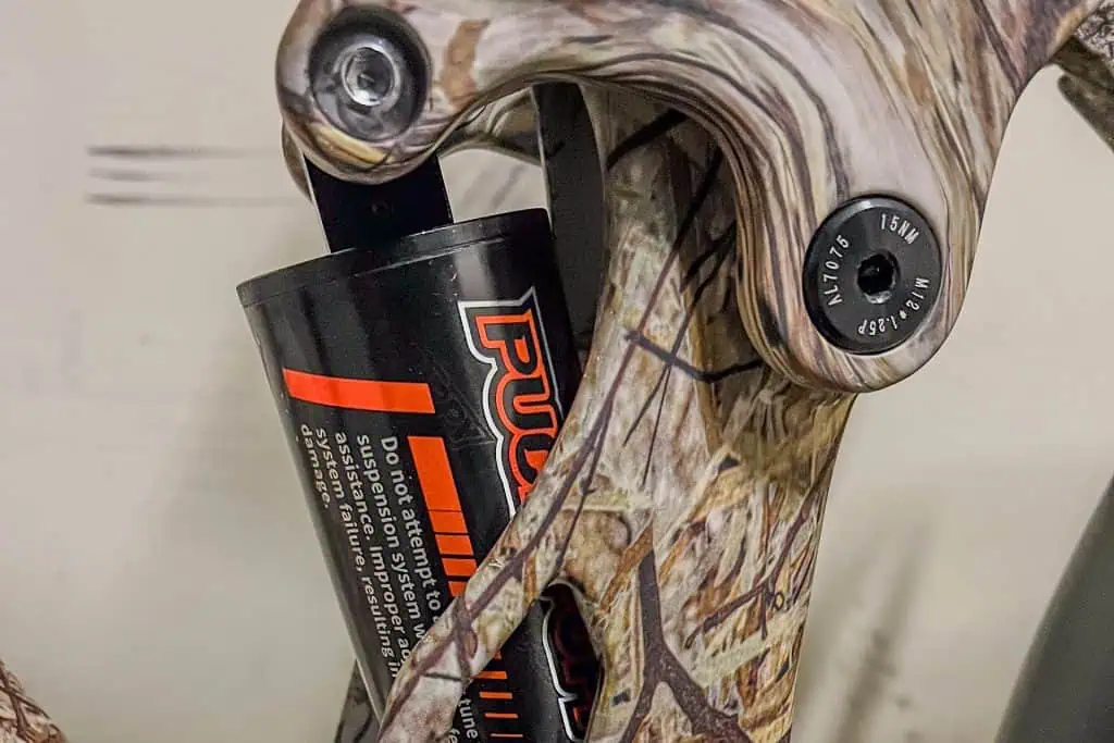Close-up of a PUCKIPUPPY Bulldog camouflage-patterned mountain bike frame, showing part of a rear suspension shock absorber with warning text and an adjustment bolt marked “15Nm MAX LOAD.”.