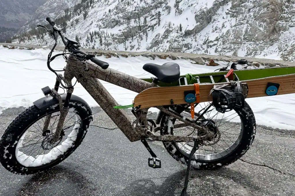 A PUCKIPUPPY Bulldog fat-tire bike with a camouflage frame is parked on a snowy mountain road, carrying a wooden splitboard on its side. Snow-covered mountains and trees create a stunning winter backdrop.