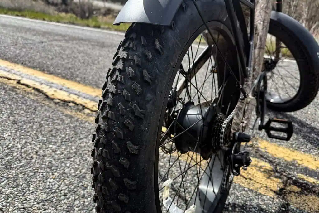 Close-up of a PUCKIPUPPY Bulldog fat-tire bicycle wheel on an asphalt road with yellow lines. The bike is partially visible, showing detailed tread and gear mechanism against a blurred backdrop of roadside vegetation.