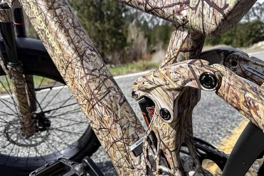 Close-up of a PUCKIPUPPY Bulldog bicycle frame with a camouflage pattern, showcasing the suspension and part of the front tire, parked on a road lined with trees and blurred greenery in the background.