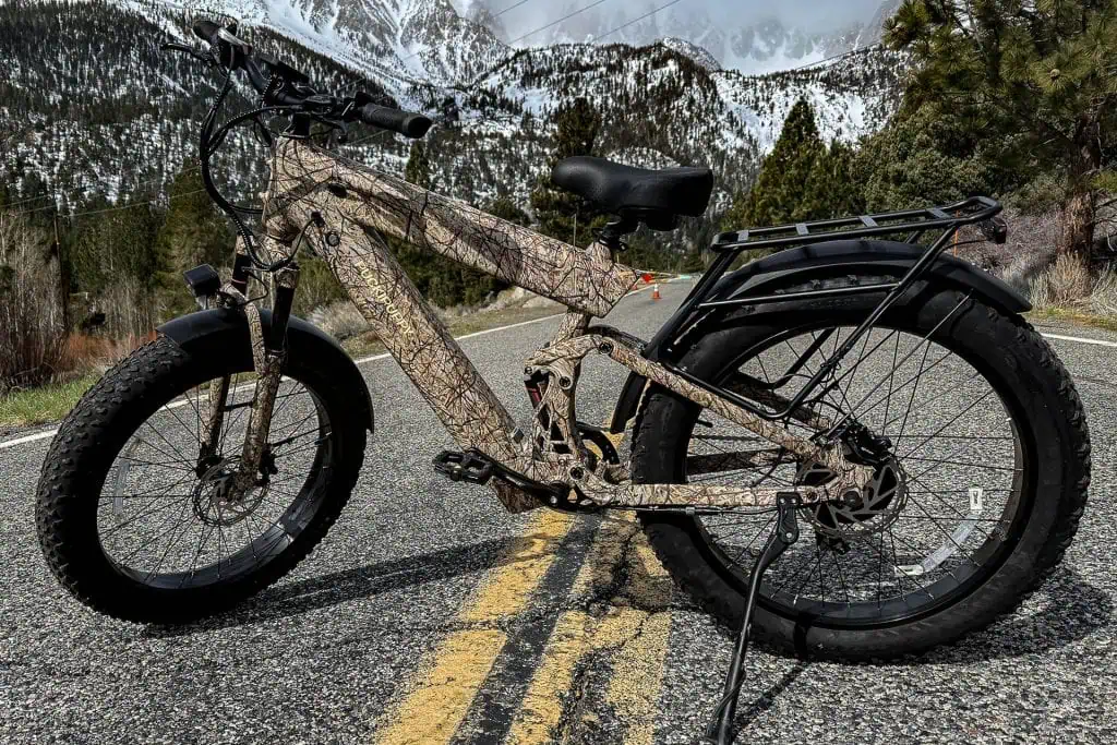 A PUCKIPUPPY Bulldog camouflage-patterned fat tire electric bike is parked on a mountain road, with snow-covered peaks, pine trees, and cloudy skies in the background.