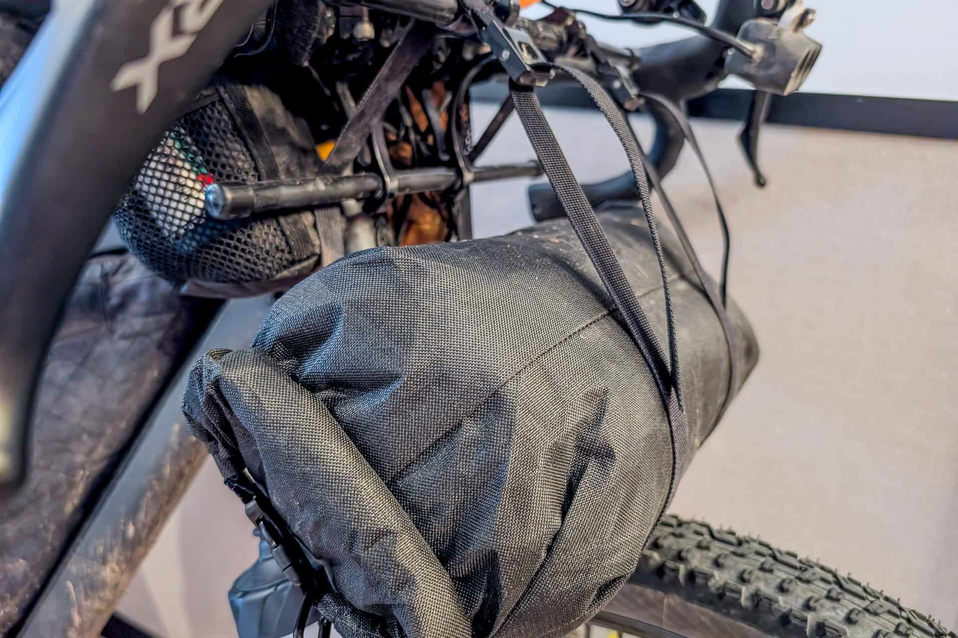 A close-up of a black Gossamer Gear Grit 28 bikepacking bag strapped to the handlebars of a bicycle, with a knobby front tire visible below. The cylindrical bag is secured firmly with adjustable straps.