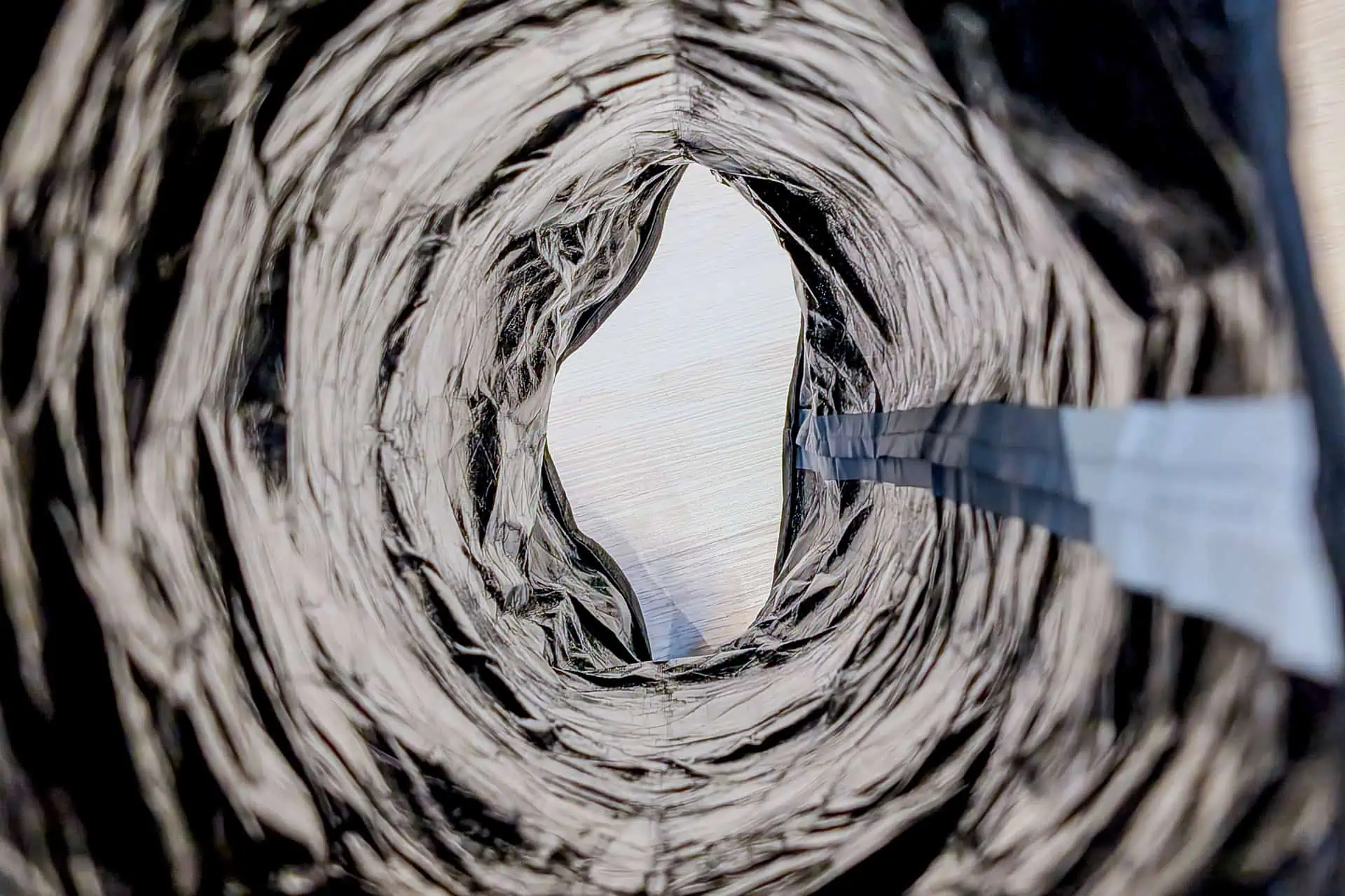 Close-up view looking through a rough, circular tunnel with textured black and gray materials, opening to a bright sky. A blue-gray strip runs along the right side, echoing the minimalist style of the Gossamer Gear Grit 28.