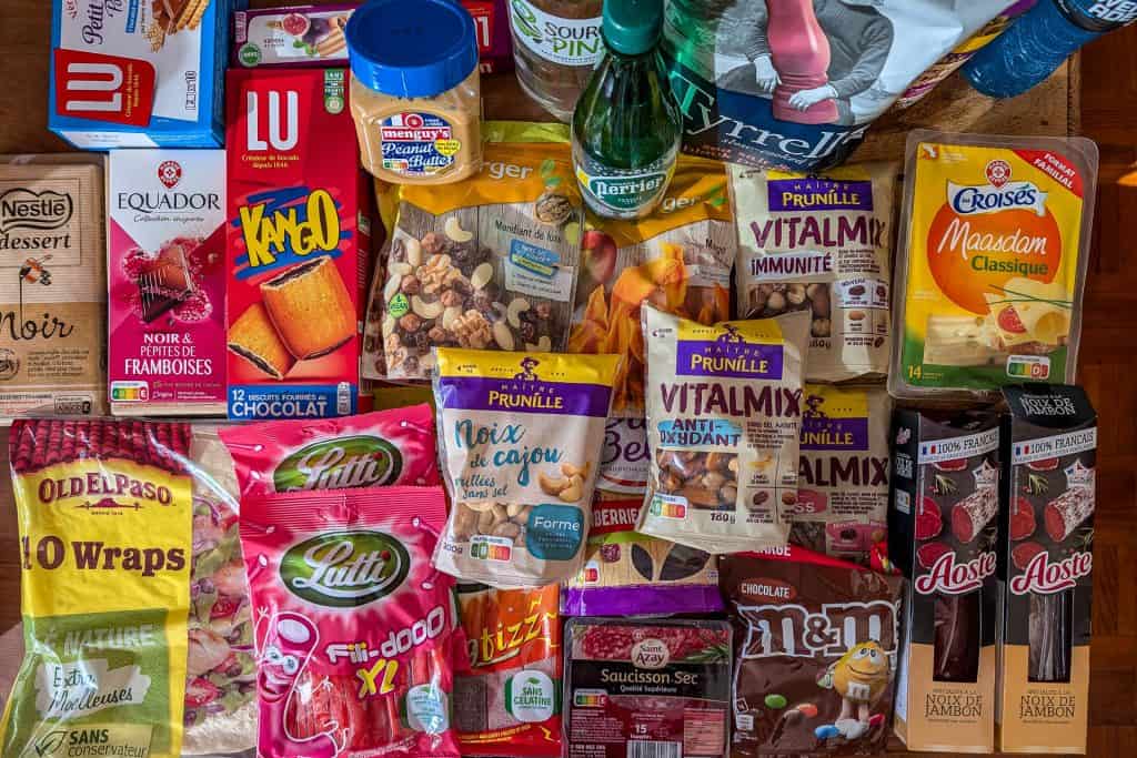 A variety of packaged snacks and food items, including crackers, chocolate, mixed nuts, juice, sparkling water, chips, candy, and dried sausage, are arranged on a table in a colorful, overlapping display.