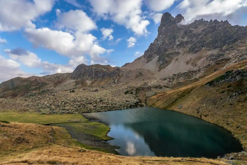A small, clear mountain lake sits at the base of rocky, rugged peaks under a partly cloudy sky. The landscape features brown grassy slopes and scattered rocks surrounding the water.