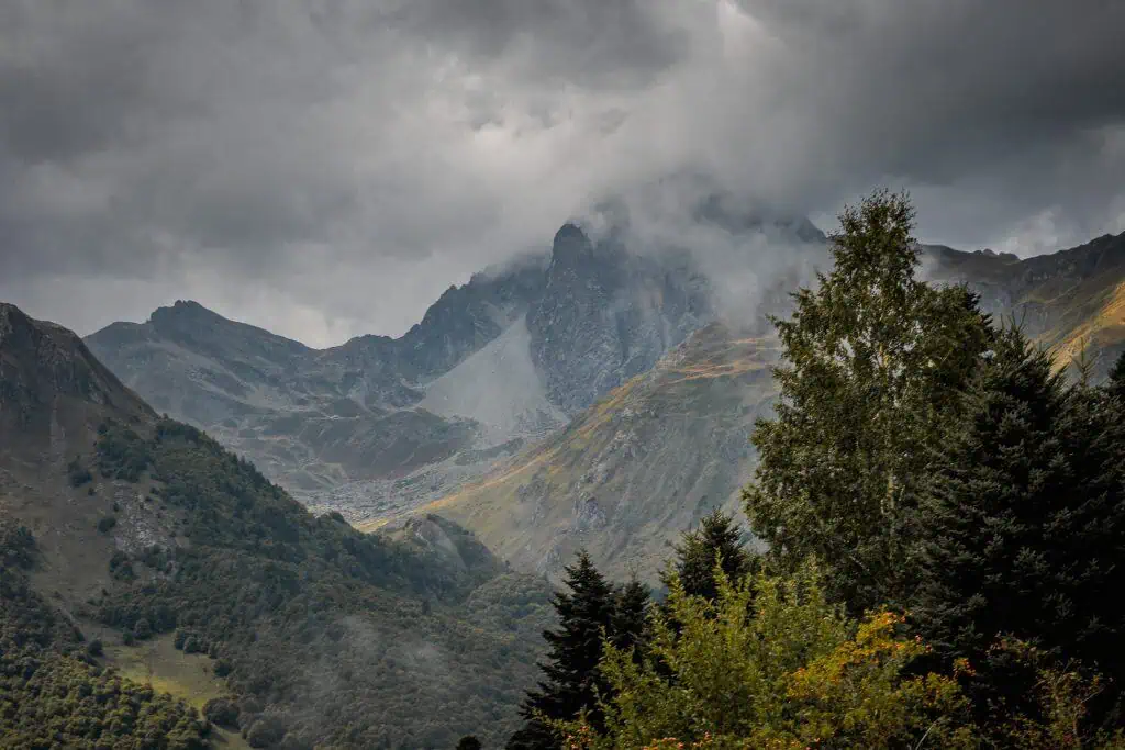 Dramatic, cloudy sky over rugged, misty mountains with steep slopes and rocky peaks; lush green trees and foliage fill the foreground.