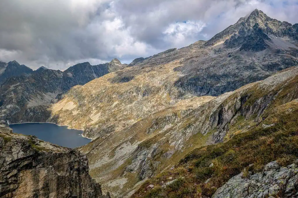 Rocky mountain landscape with a clear blue lake nestled at the base, surrounded by rugged peaks along the Pyrenees High Route (HRP) under a partly cloudy sky. Sparse vegetation clings to the steep terrain in the foreground—perfect for any photo gallery.