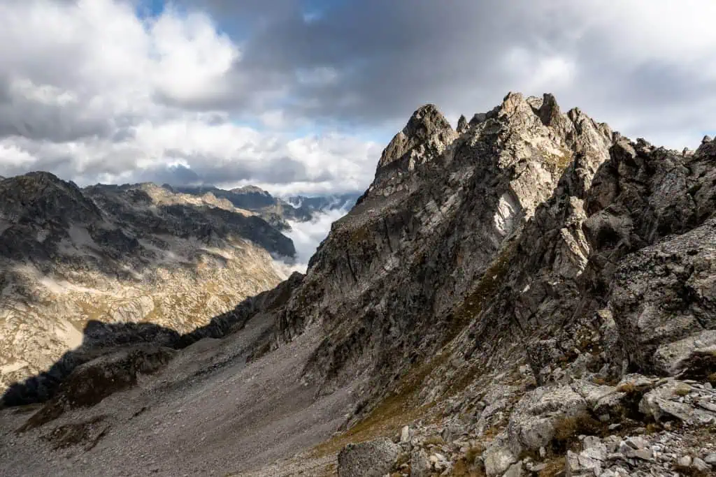 Rocky mountain peaks rise beneath a dramatic sky with clouds. Sunlight highlights the rugged slopes of the Pyrenees High Route, while mist and distant mountains fill the background, creating a striking alpine landscape for any photo gallery.