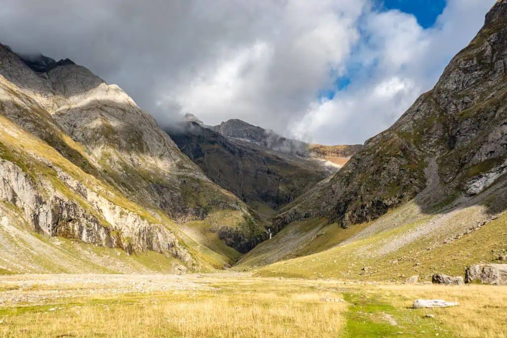 A wide, grassy valley nestled between steep, rocky mountains under a partly cloudy sky—an iconic scene along the Pyrenees High Route. Sunlight highlights patches of grass and rugged slopes, while misty clouds hang over the distant peaks.