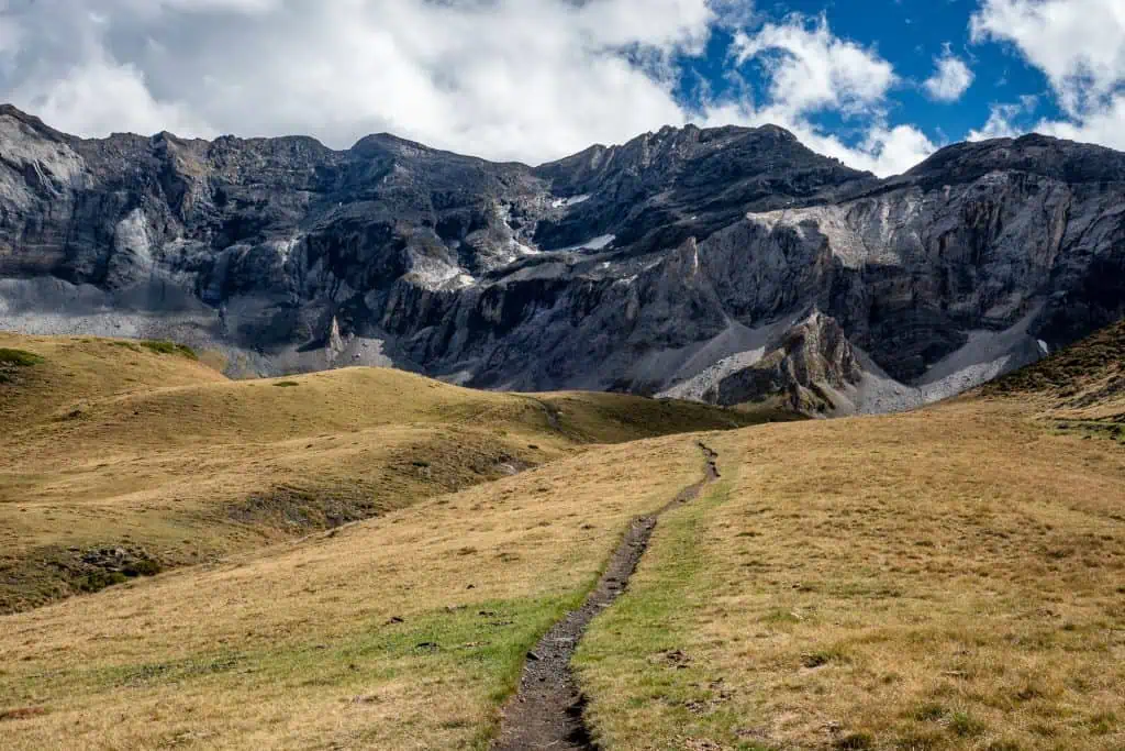 A narrow dirt trail winds through a grassy meadow toward rugged, rocky mountains under a partly cloudy sky—an inviting scene along the Pyrenees High Route (HRP), perfect for any adventure photo gallery.