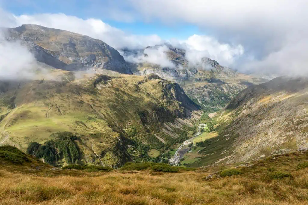 A wide valley with green grass, scattered trees, and rocky slopes, surrounded by the tall, rugged mountains of the Pyrenees High Route (HRP), partially covered by clouds under a bright sky. A river winds through the valley below.