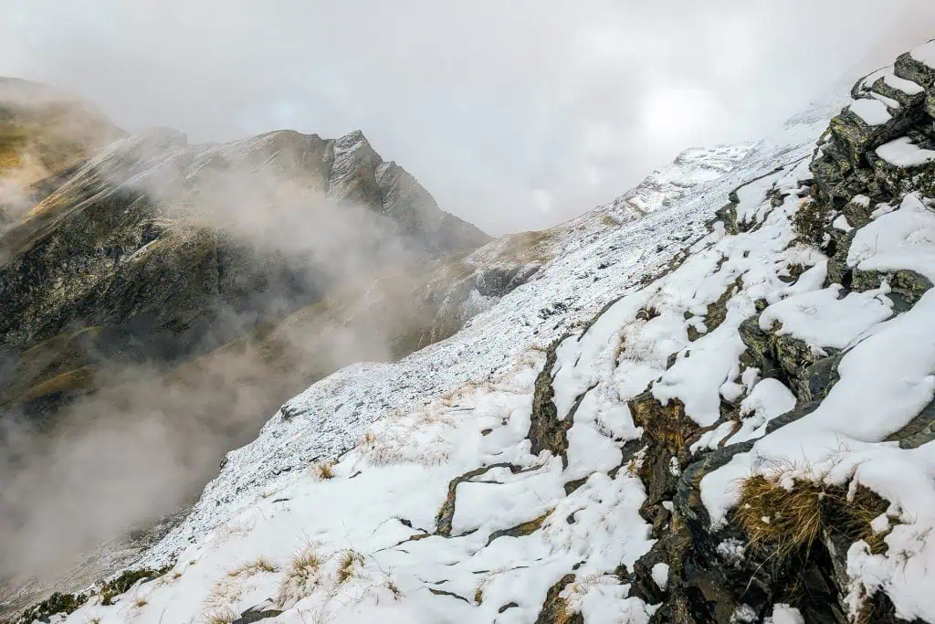 Snow-covered mountain slope with rocky outcrops and patches of grass, shrouded in mist and clouds along the Pyrenees High Route (HRP), with rugged peaks in the background under a cloudy sky—perfect for any photo gallery.