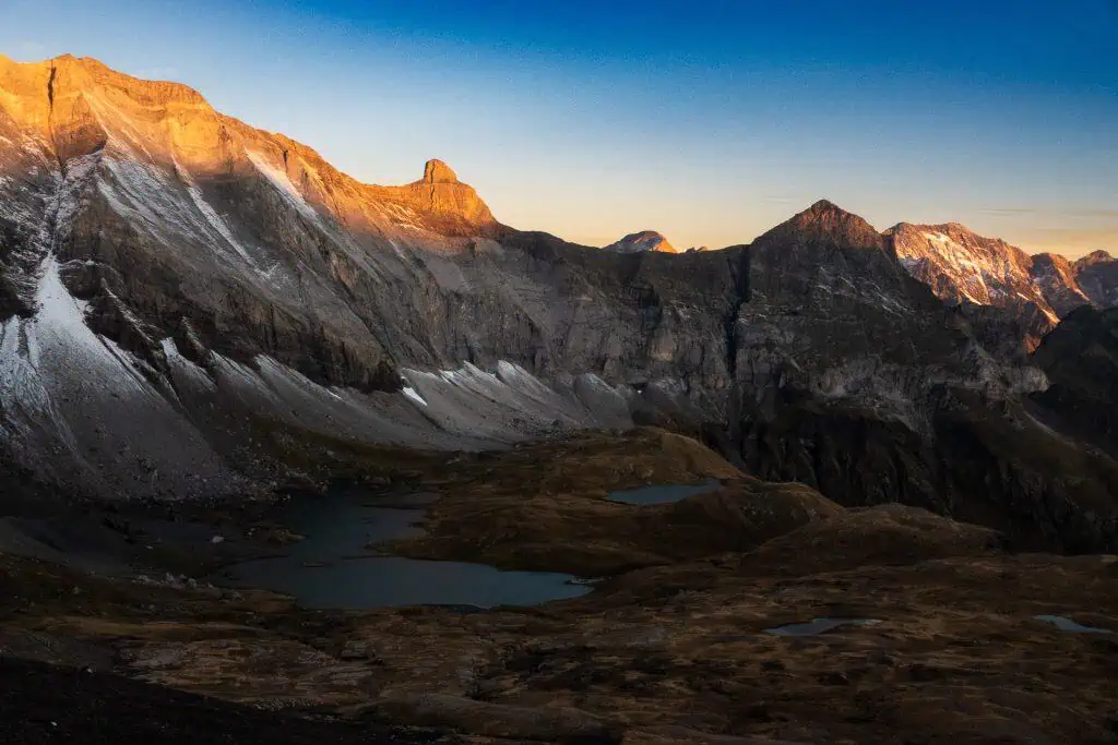 Sunlight illuminates the peaks of rugged, rocky mountains along the Pyrenees High Route. Below, alpine lakes and brown grass fill the valley, while the sky glows with soft blue and golden hues at sunrise or sunset.