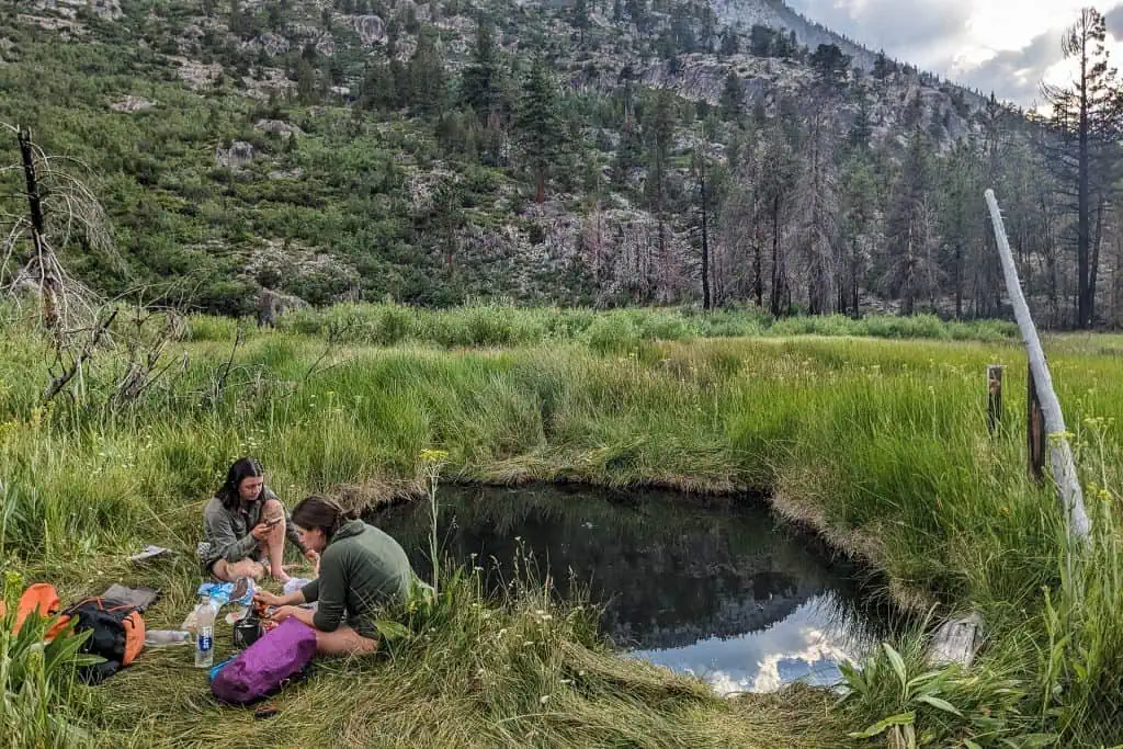 Two people sit on grass by a small pond surrounded by tall green grass, backpacks and supplies nearby—a moment of rest during their JMT hikes, with forested mountains and a mostly cloudy sky in the background.