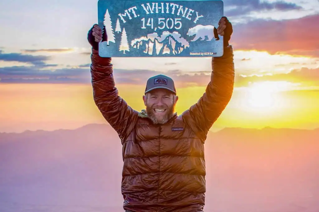 A smiling man in a puffy jacket and cap holds up a Mt. Whitney 14,505 sign at sunrise, mountains and a colorful sky behind him—celebrating the finish after the JMT.