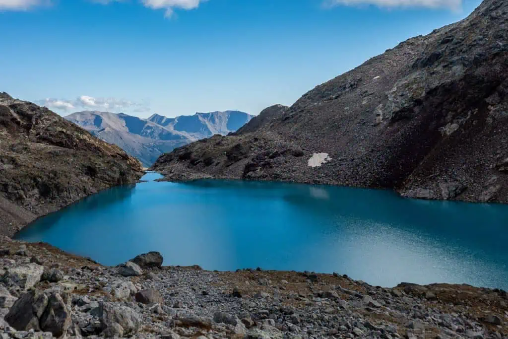 A vibrant blue alpine lake along the Pyrenees High Route (HRP), surrounded by rocky, barren mountains, with distant peaks visible under a clear blue sky—a perfect addition to any photo gallery.