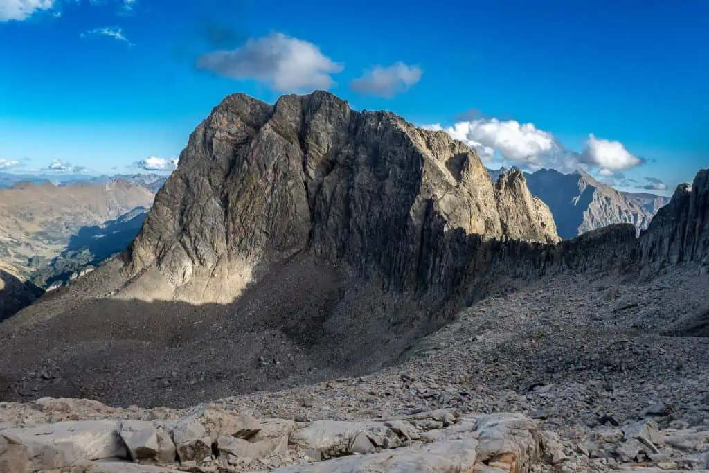 A rugged, rocky mountain peak rises sharply against a blue sky with scattered clouds along the Pyrenees High Route. Sunlight and shadows highlight the steep slopes, while the foreground is covered with loose rocks and stones.