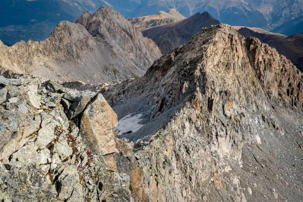 A rugged mountain ridge with jagged rocky peaks and steep slopes, viewed from a high vantage point along the Pyrenees High Route, with distant mountains visible in the background under clear daylight.