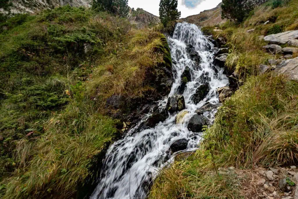 A small waterfall cascades over rocks surrounded by lush green grass and vegetation on a sloped hillside under a partly cloudy sky, capturing the natural beauty along the Pyrenees High Route for your Photo Gallery.