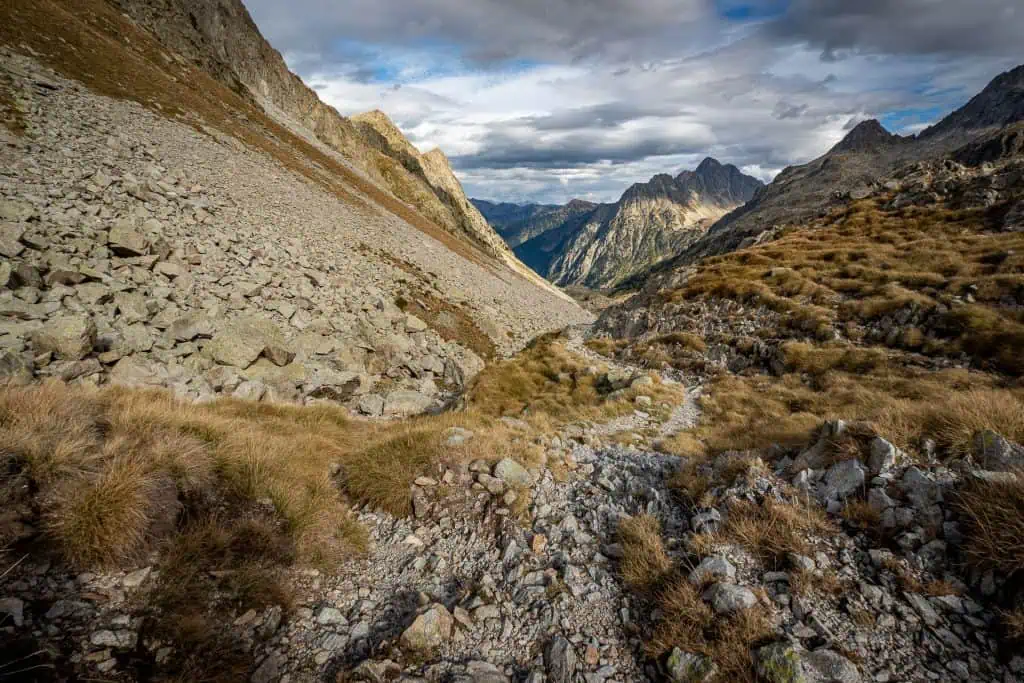 Rocky mountain landscape along the Pyrenees High Route, with a narrow, stony trail winding through grassy slopes and rugged terrain, leading toward distant peaks under a partly cloudy sky—perfect for an HRP photo gallery.
