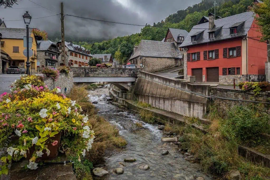A stream runs under a bridge in a small village with stone and red buildings, surrounded by trees and mountains along the Pyrenees High Route; colorful flowers decorate the foreground and cloudy skies loom overhead.