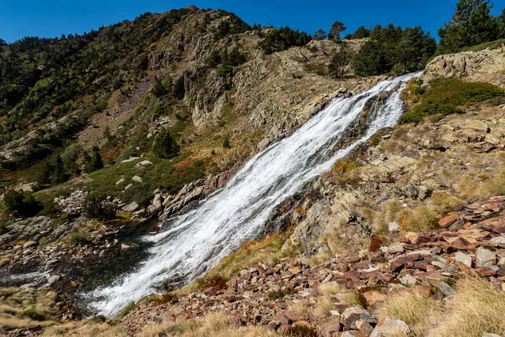 A waterfall cascades down a rocky mountainside along the Pyrenees High Route, surrounded by sparse grass, shrubs, and pine trees under a clear blue sky.
