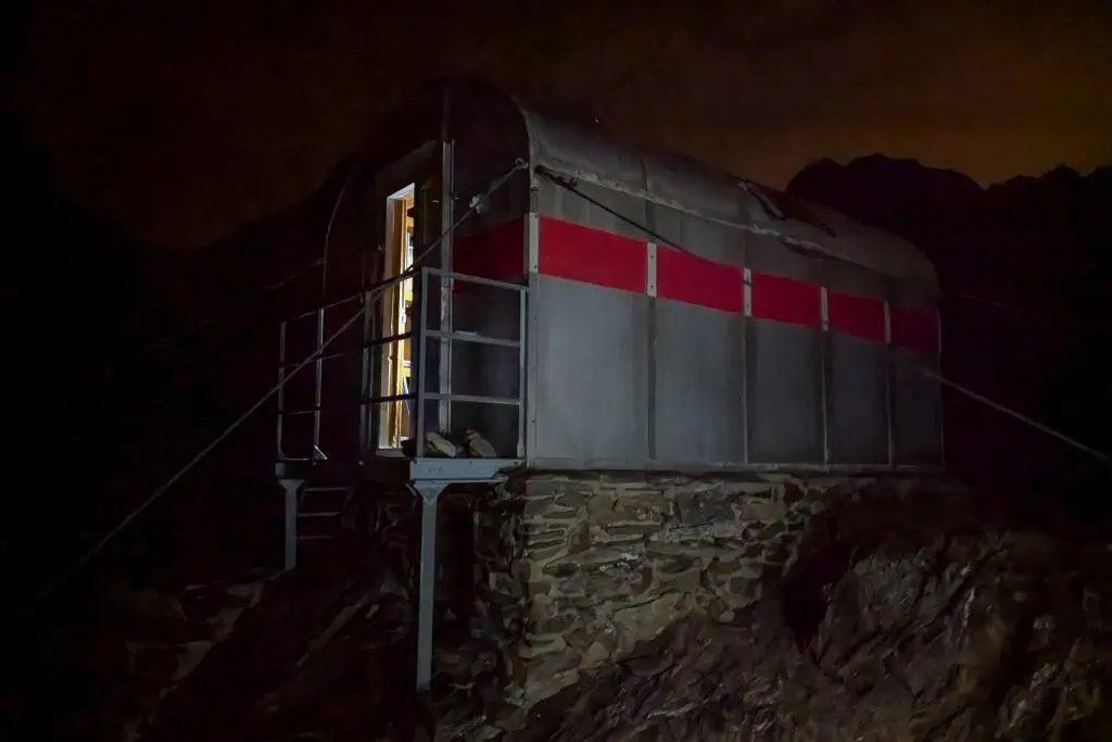 A small metal mountain hut with a red stripe, elevated on a stone base, is dimly lit from within at night along the Pyrenees High Route, surrounded by darkness and rocky terrain—perfect for any HRP photo gallery.