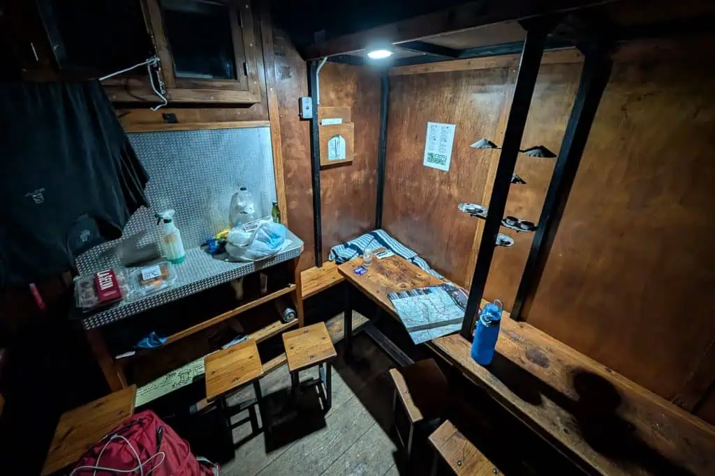 A small, rustic wooden room along the Pyrenees High Route with benches, stools, a table holding a map, water bottle, and headlamp. Kitchen area has a sink, dishes, and jug. Clothes hang above; climbing gear and an HRP photo gallery are on the wall.